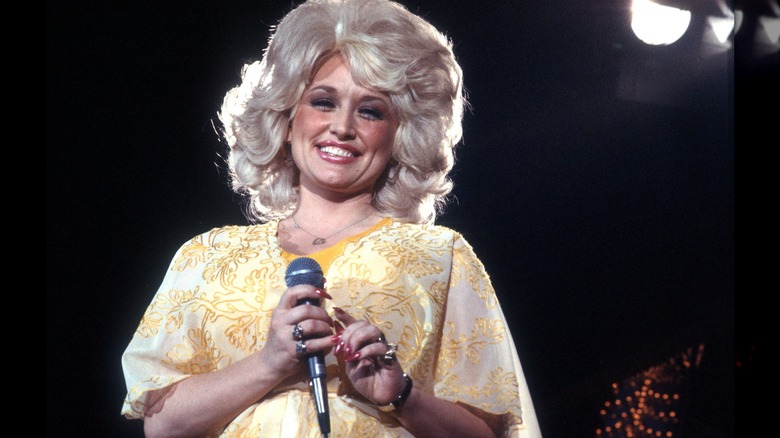 Dolly Parton in yellow with red nails while holding a microphone