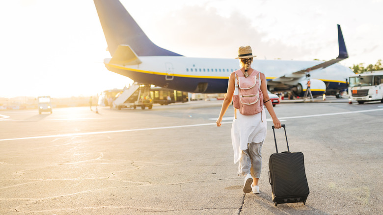 A woman walking towards a plane pulling a suitcase and carrying a pink backpack
