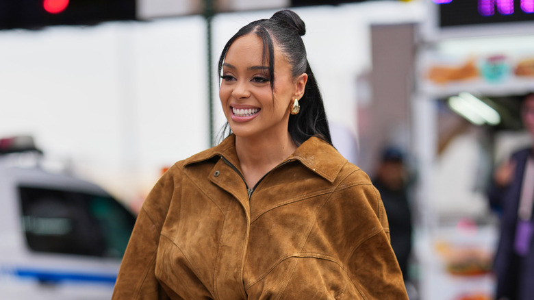 A woman walking in a brown jacket with face framing tendrils and a half up bun