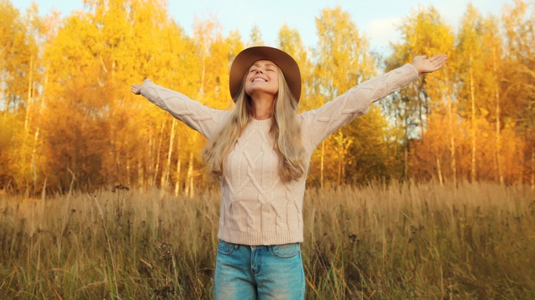 A woman wearing a sweater, jeans, and a hat while reaching her arms out in front of a row of trees in fall