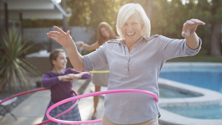 An older woman hula hooping by a swimming pool with her daughter and granddaughter in the background