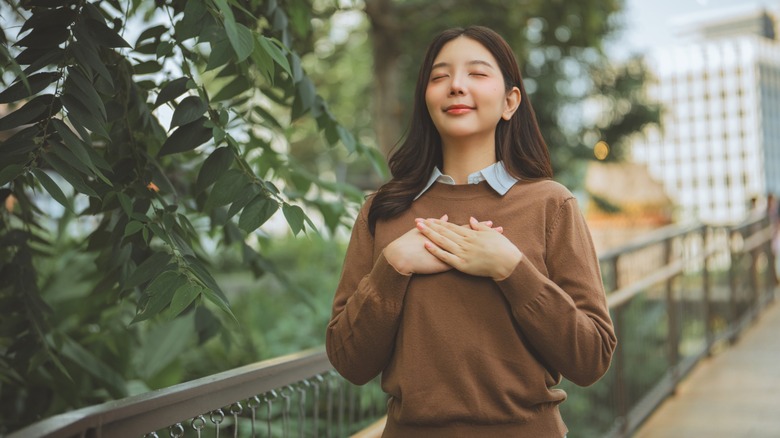 A woman standing outside with her eyes closed and her hands on her chest