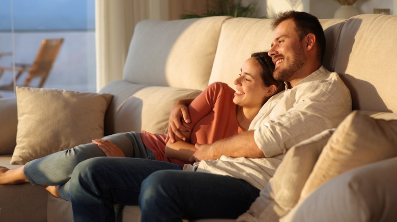 A couple sitting on the sofa together and smiling, with the woman's head resting on the man's shoulder