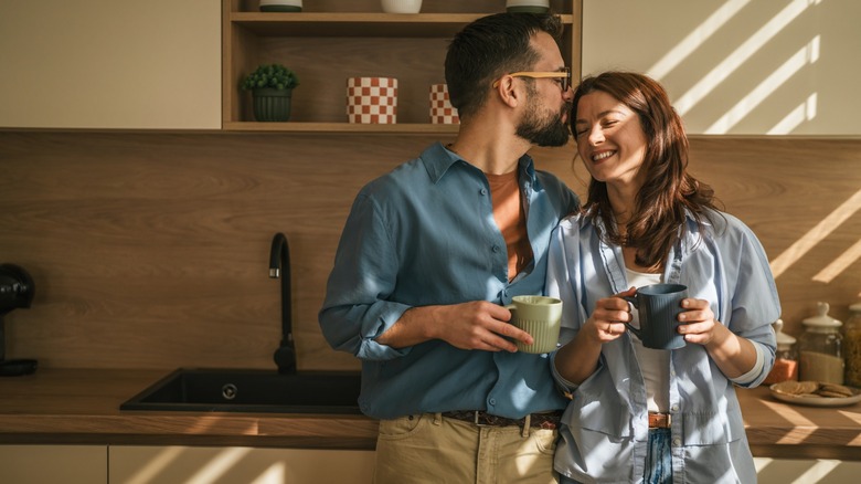 A man planting a kiss on a woman's forehead while they hold mugs in a kitchen