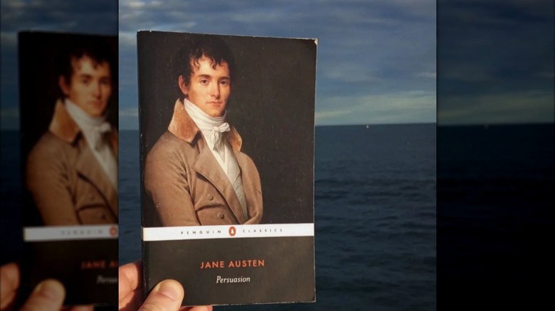 A hand holding a copy of "Persuasion" in front of the ocean