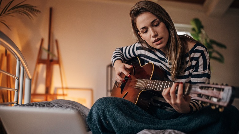 A woman playing the guitar while sitting on her bed