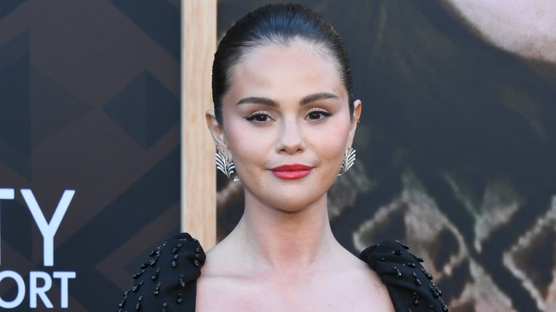 Selena Gomez smiling on the red carpet in a black dress while posing in front of a black background