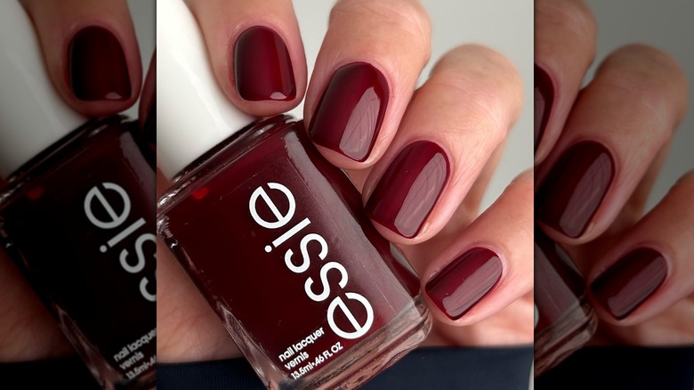 A close-up of a person with Essie's bordeaux shade painted on their nails, holding the nail polish bottle