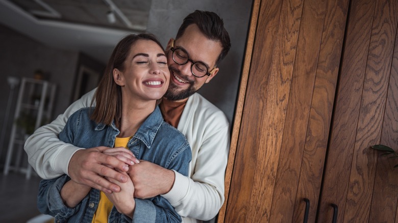 A couple smiling while the man hugs the woman from behind