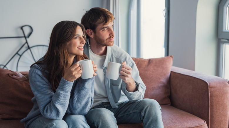 A couple drinking from mugs while looking out a window