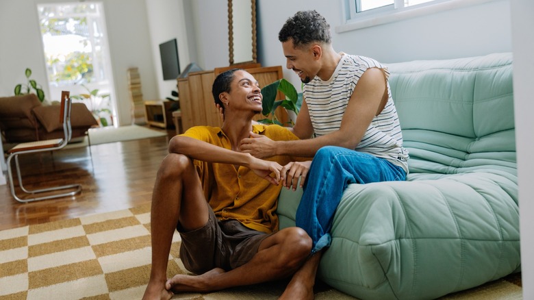 A couple smiling at one another while one sits on a sofa and the other on the floor