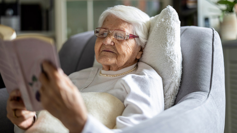 An elderly woman rests comfortably on a chair as she reads a book