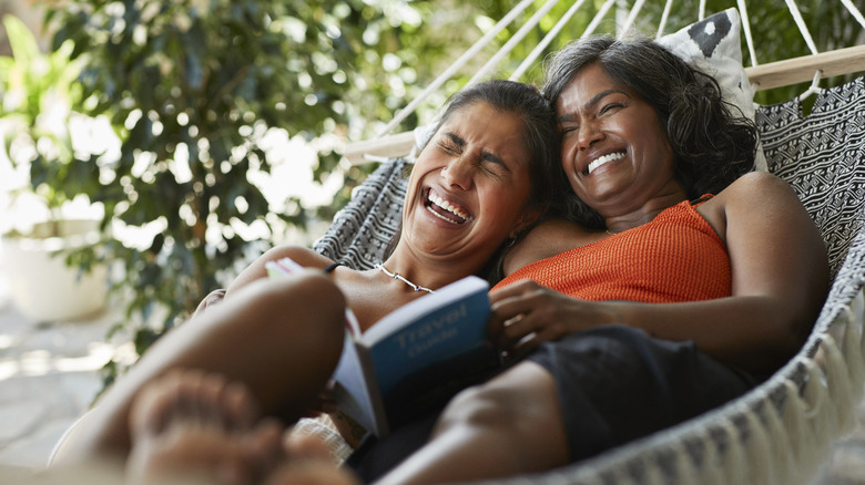 Two women share a hammock and laugh while holding a book