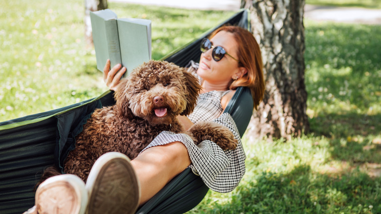 A woman lying in a hammock with a dog and reading a book