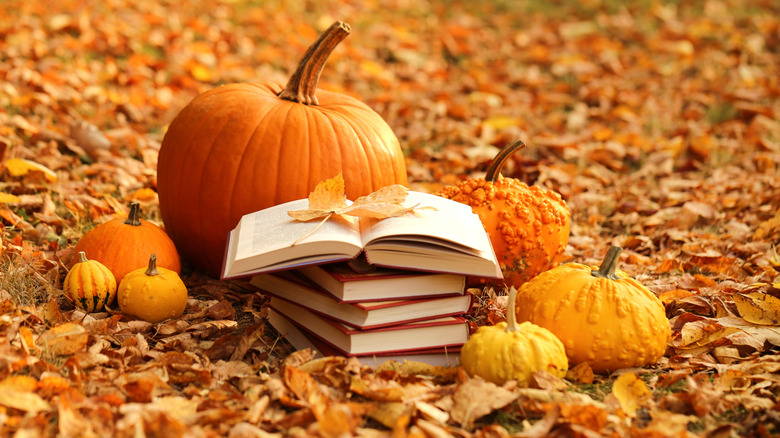 Open book placed atop a stack of books and surronded by fall leaves and pumpkins