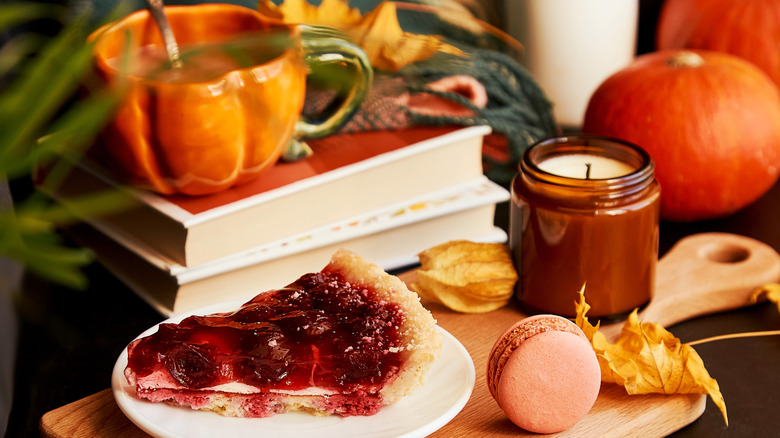 Stack of books, with a pumpkin-shaped mug on top, sitting next to a slice of pie, candle, and macaron