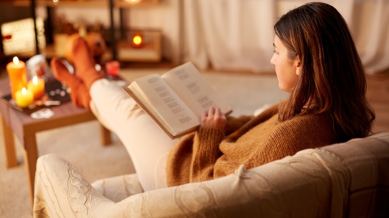 Lady reads a book in blanket on the sofa in front candles
