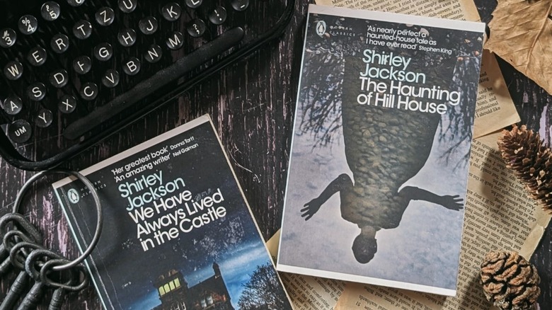 Two hard cover edition of Shirley Jackson books, including "The Haunting of Hill House" styled next to a typewriter, vintage-looked paper, a set of iron kets, and pine cones.