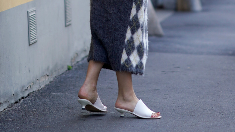 Side view of white kitten heels featuring one, large strap across the foot being worn with a gray and plaid skirt
