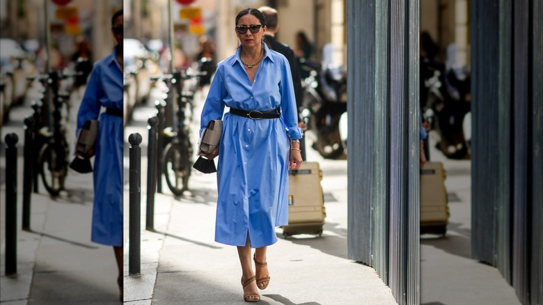 A woman in a blue shirt dress walking outside with sunglasses and a black belt