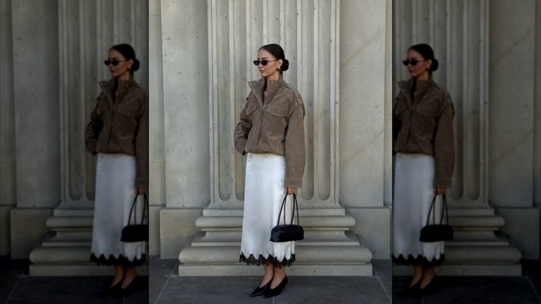 A woman standing in front of a marble building in a suede jacket and white slip skirt