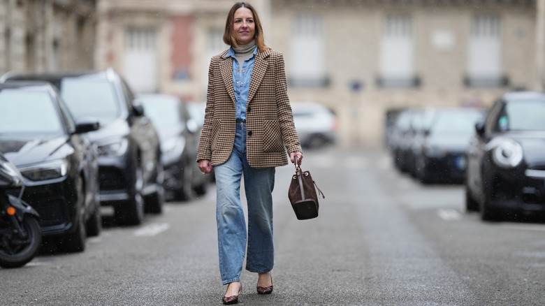 Alba Garavito Torre wears long light brown hair worn loose with a center part, natural makeup with defined brows and soft lipstick, a beige turtleneck pullover, a light blue denim shirt layered, a beige and brown houndstooth pattern blazer jacket