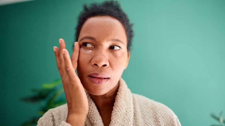 A woman in a bathrobe in front of a green background applying eye cream and looking to the side