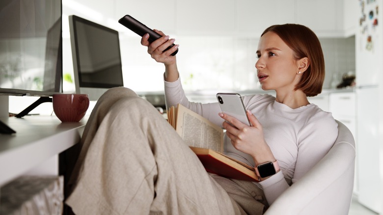 A woman sitting down while holding a book, TV remote and her cell phone