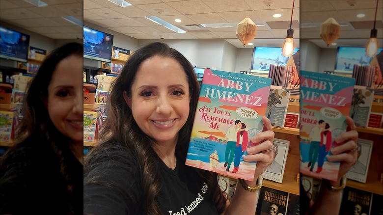 Abby Jimenez smiling for a selfie while holding up her book "Say You'll Remember Me"