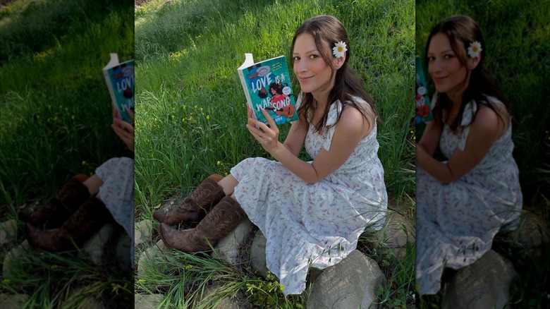 Danica Nava sitting on a rock, smiling at the camera while holding her book "Love is a War Song" open