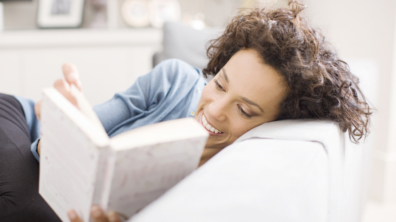 A woman lies on the couch comfortably and smiles as she reads a book