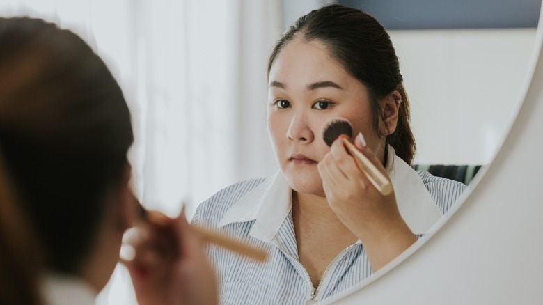 A woman with her dark hair tied back applying blush to her cheeks in the mirror