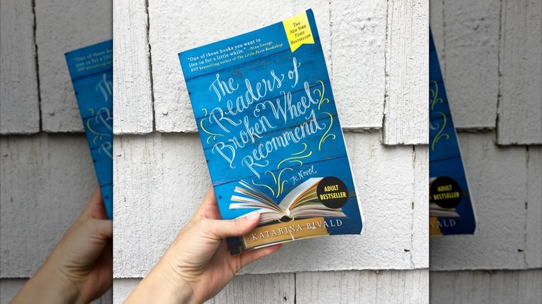 A hand holding up a copy of "The Readers of Broken Wheel Recommend" in front of the side of a building