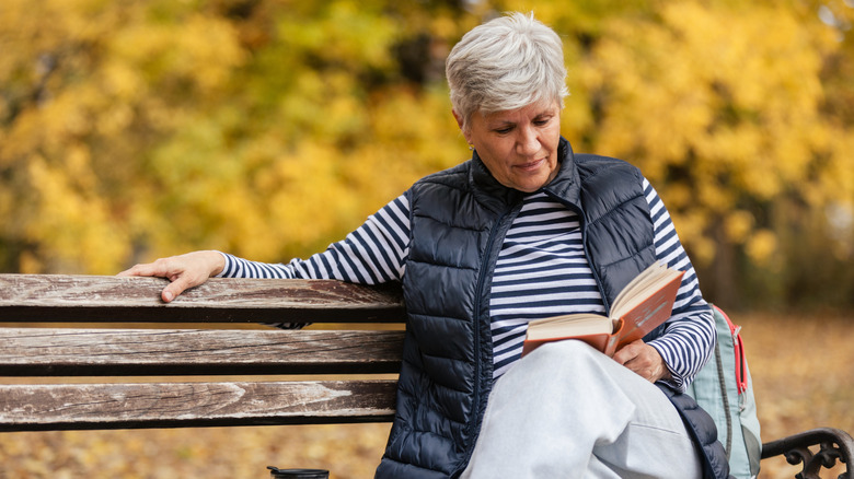 Senior woman relaxing on a park bench, reading a book during autumn