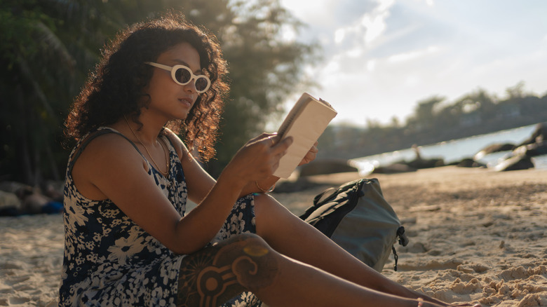 Curly-haired woman with white sunglasses sitting on the beach while reading an open book