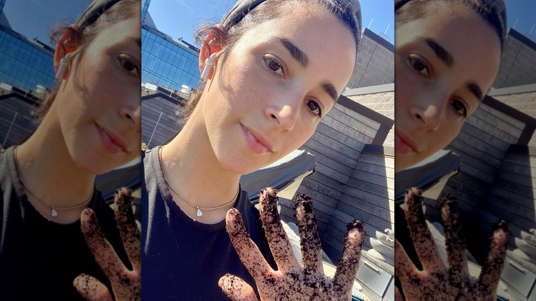 Aly Raisman with her hair tied back and a wireless earphone in her ear holding up a hand with dirt on it