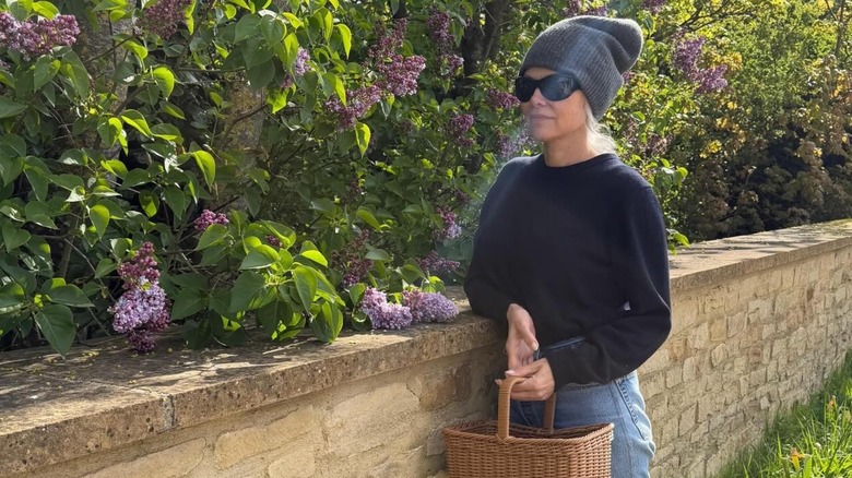 Pamela Anderson leaning against a stonewall with healthy foliage and purple flowers in the background while holding a basket