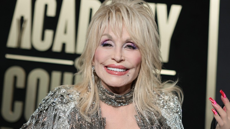 Dolly Parton smiling wearing colorful makeup in front of a black and gold background