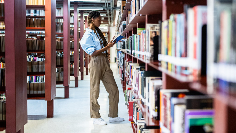Long shot of woman with a braidedhairstyle and khaki slacks browsing books in a library