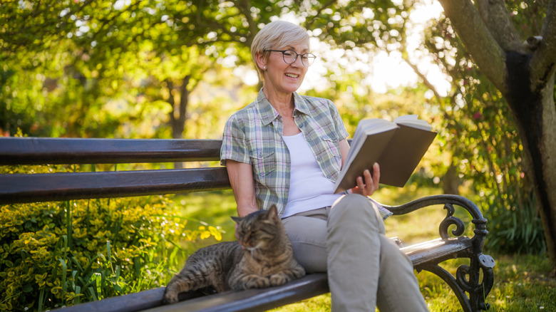 Happy senior woman enjoys reading book and spending time with her cat while sitting on a bench in her garden.