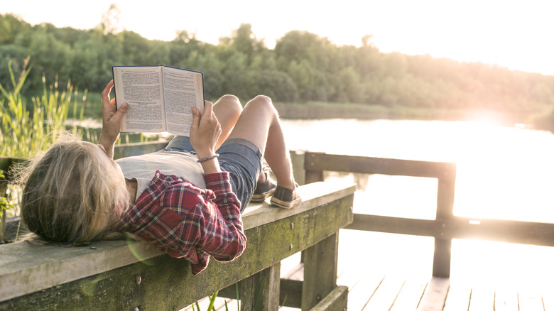 oung girl reading a book while lying on her back on a wooden beam at a wooden dock at the lake