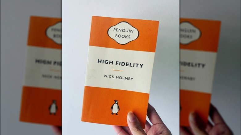 A hand holding up a copy of "High Fidelity" in front of a white wall