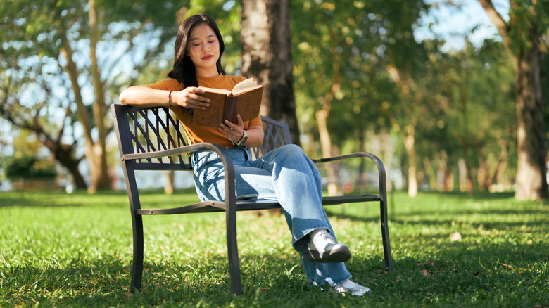 A woman sitting on a park bench reading a book with her legs crossed