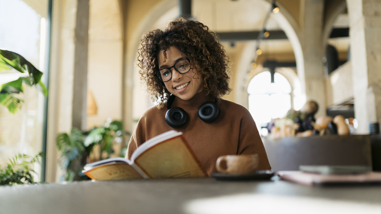 A woman sitting at a table with a cup of coffee while reading and book and wearing headphones around her neck