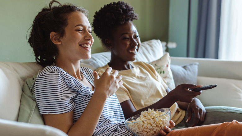 Two women sit comfortably on a couch with a bowl of popcorn watching TV