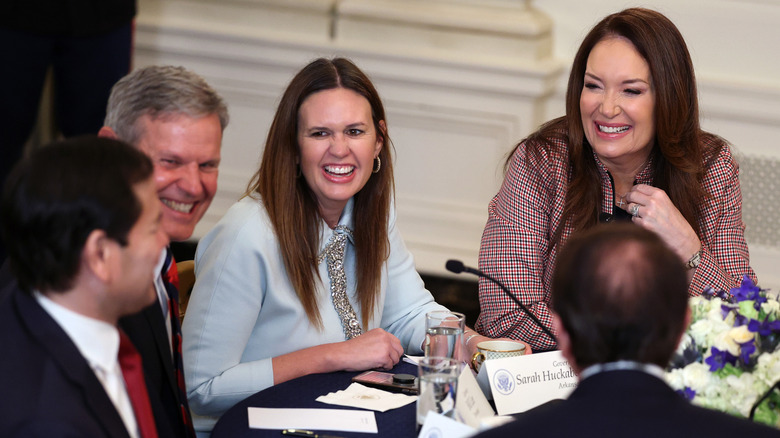 Brooke Rollins and Sarah Huckabee Sanders sitting at a table and laughing with various men in suits