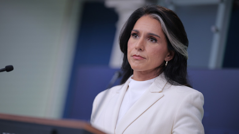 Tulsi Gabbard wearing a white suit while standing at a podium and frowning