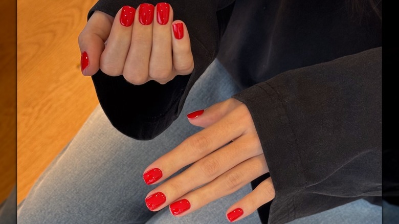 A woman's hands showing her short, red manicure while wearing a black top and blue jeans
