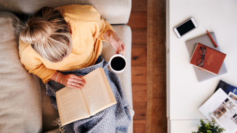 Overhead Shot Looking Down On Woman At Home Lying On Reading Book And Drinking Coffee