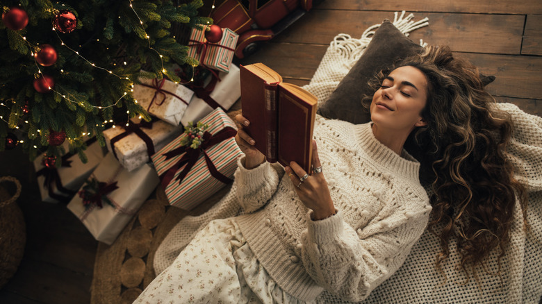 Young woman reading book, lying near Christmas tree in a cozy decorated room